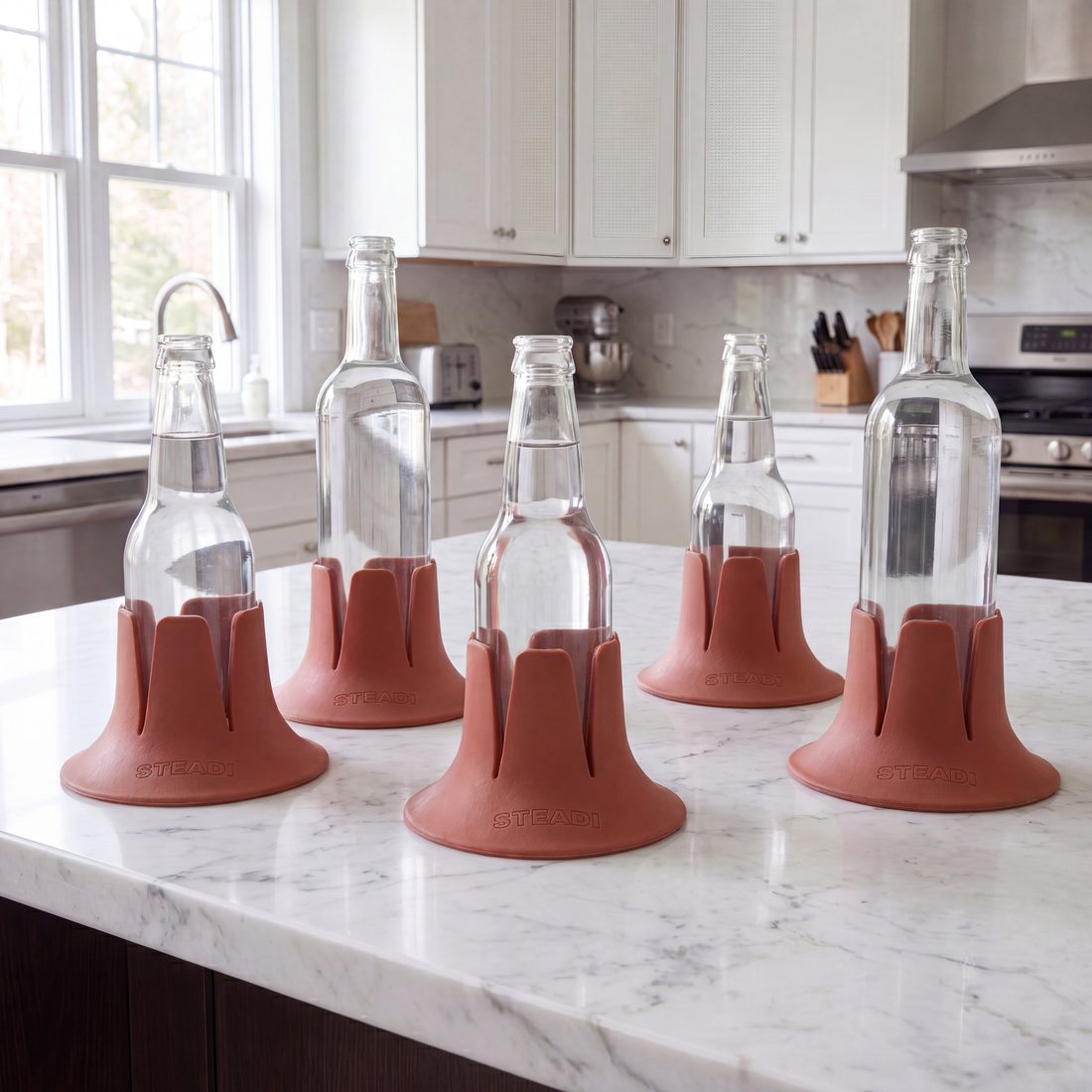 Five clear bottles of different shapes, each standing in a terracotta Steadi, along a kitchen counter.